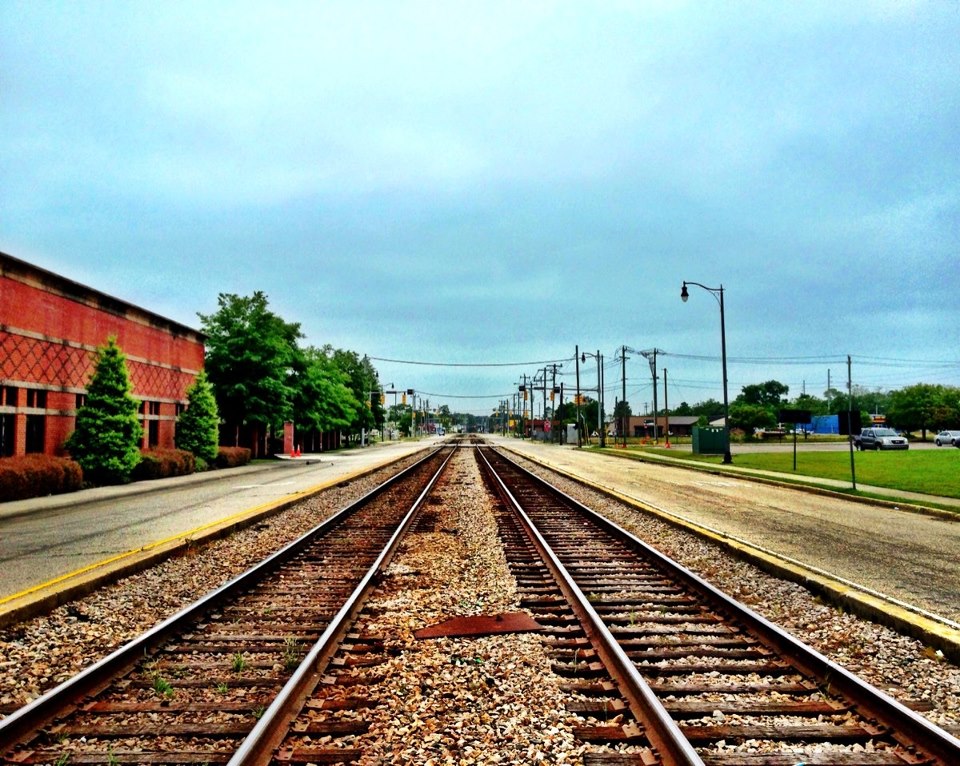 A photo showing two sets of train tracks. They converge in the distance. To their left is a red building. To the right, a bit of grass and a parking lot. Wires and trees sit in the middle distance. Above them, a light blue sky.