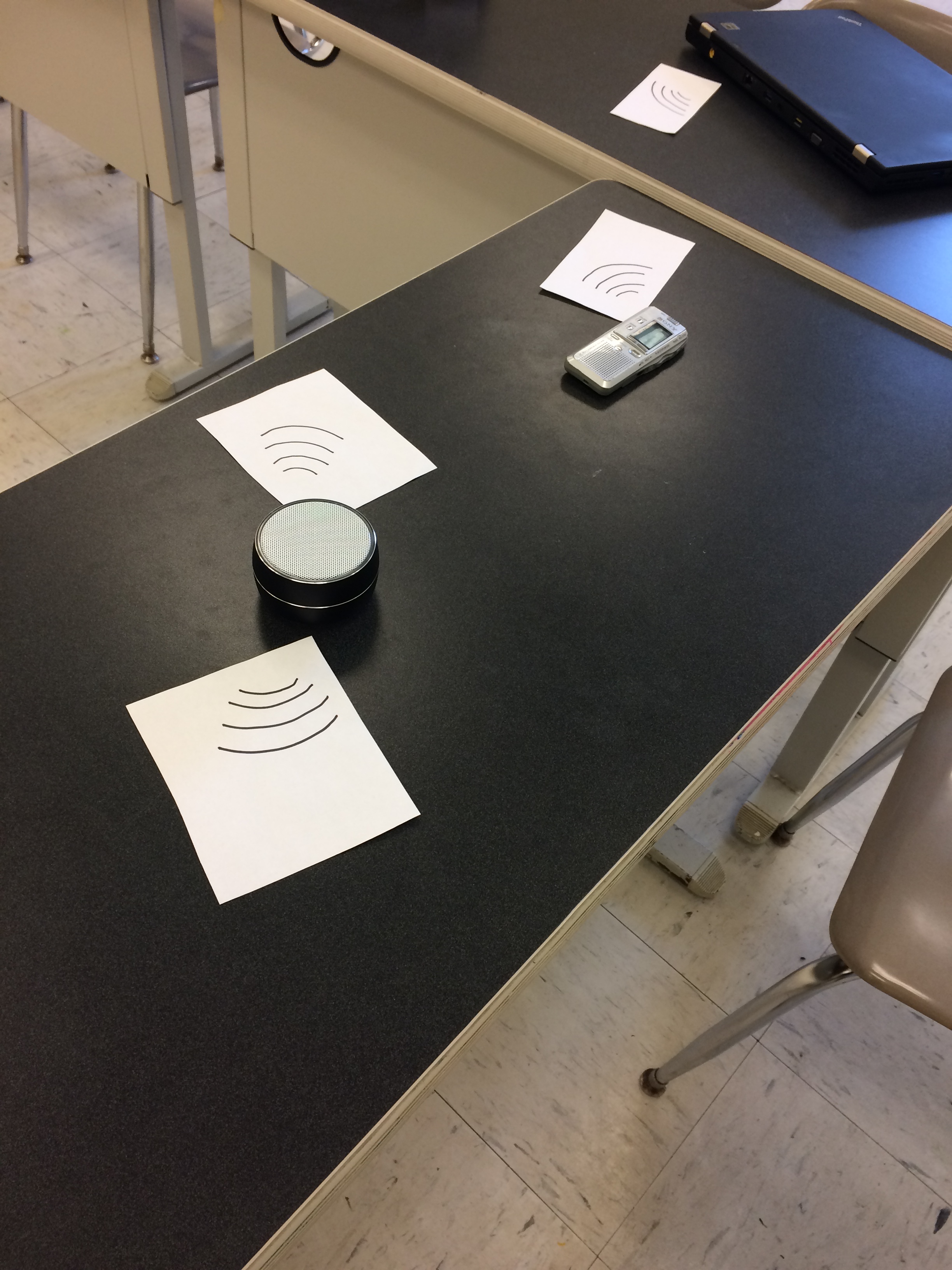 Various devices, such as a laptop and speaker act as sound sources, intentionally placed on tables within a classroom to create a sense of space.