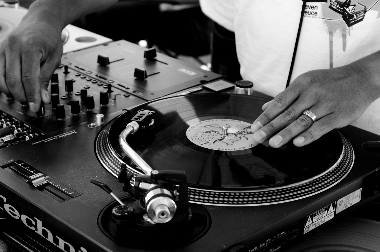 Photo of a turntable and a mixer used by a DJ. The DJ has one hand above the record and one hand on a knob on the mixer.