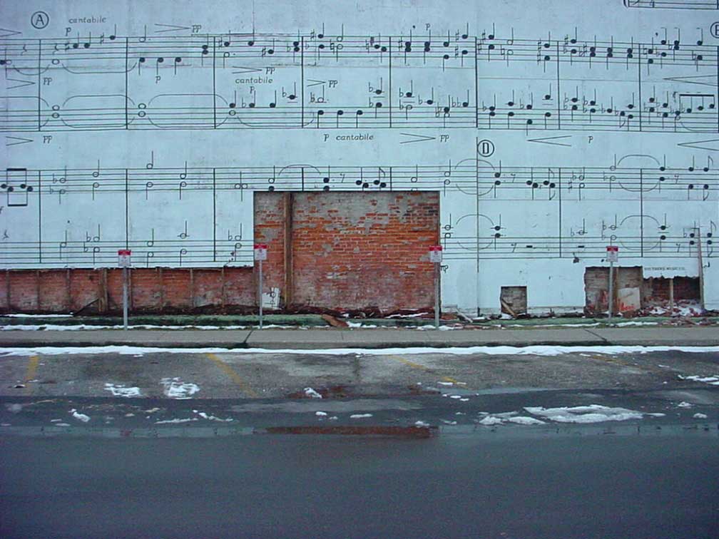 Image of a blue wall with a musical score painted on it. The bottom portion of the wall has been torn away, revealing the brick beneath. An asphalt road with parking spaces and melting snow is in the foreground.